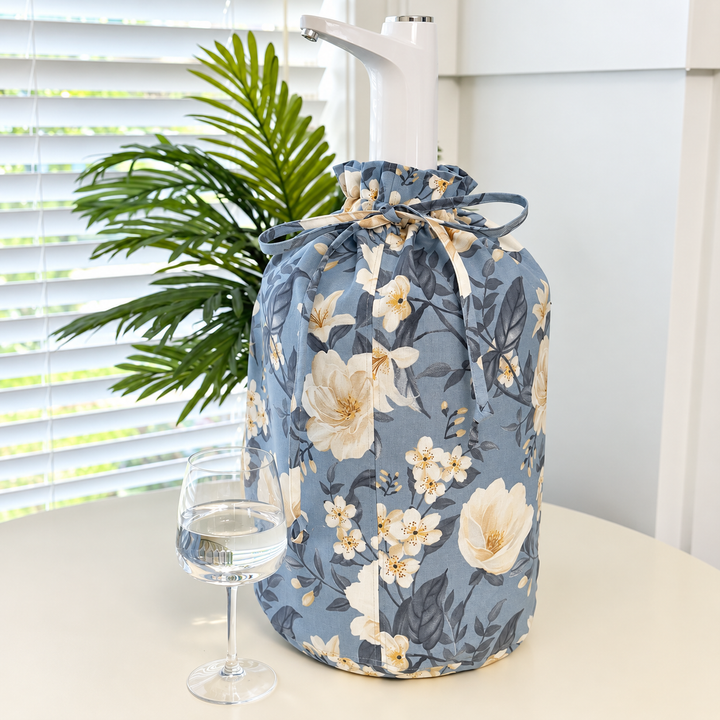 A floral drawstring bag in dusty blue with cream and white flowers, shown on a table next to a wine glass and a green plant in front of window blinds.