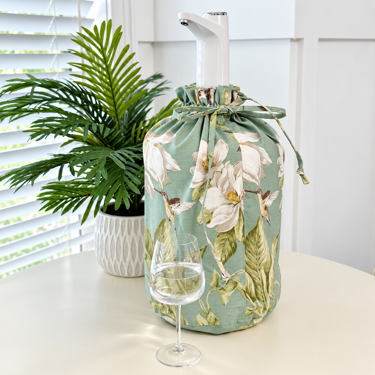 A sage green wine bottle cover with a white magnolia and hummingbird floral print, shown on a white table with a wine glass and a potted plant in a bright room.