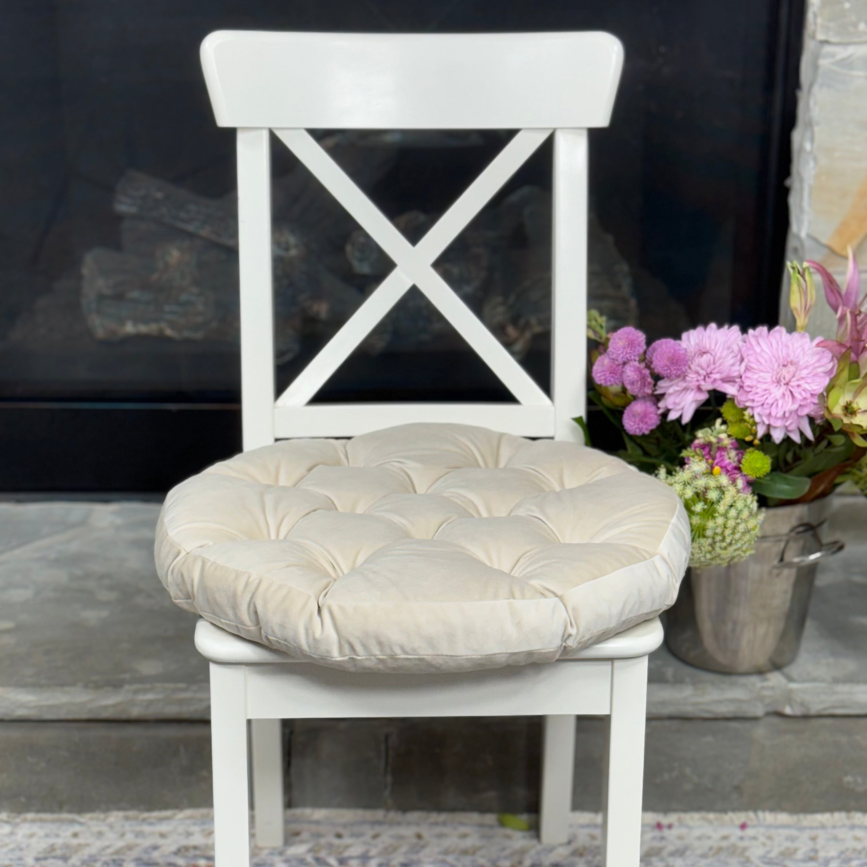 A white wooden dining chair with a tufted beige fabric seat cushion, positioned in front of a fireplace with a floral arrangement on a patterned rug.