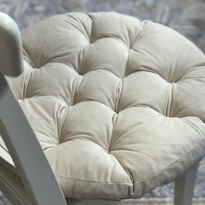 A white wooden dining chair with a large, round, tufted cream cushion on the back, styled in an indoor setting with a patterned rug and a vase of purple flowers.