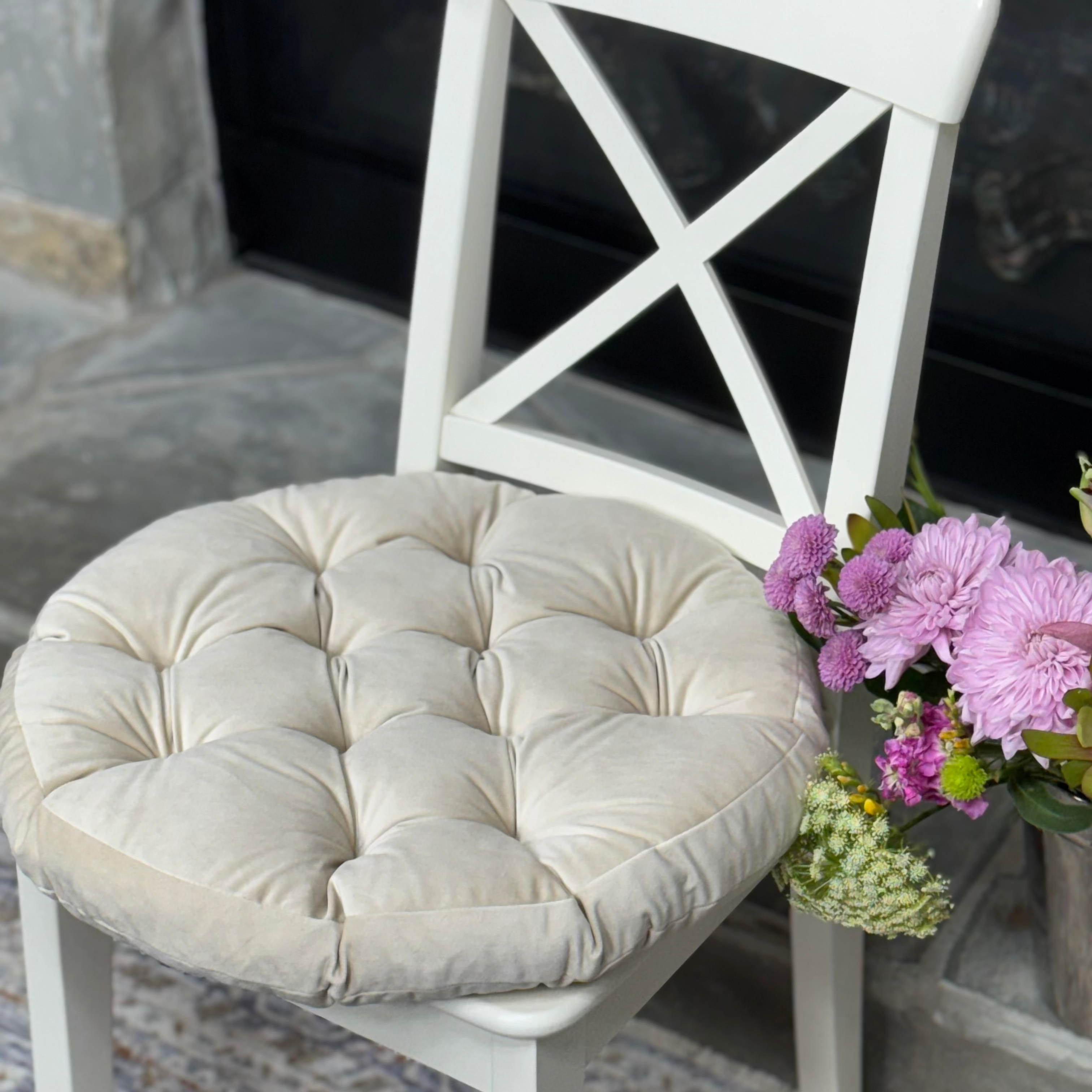 A round tufted cream velvet chair cushion on a white wooden dining chair, styled next to a vase of pink flowers on a patterned rug.