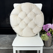 A white wooden dining chair with a large, round, tufted cream cushion on the back, styled in an indoor setting with a patterned rug and a vase of purple flowers.