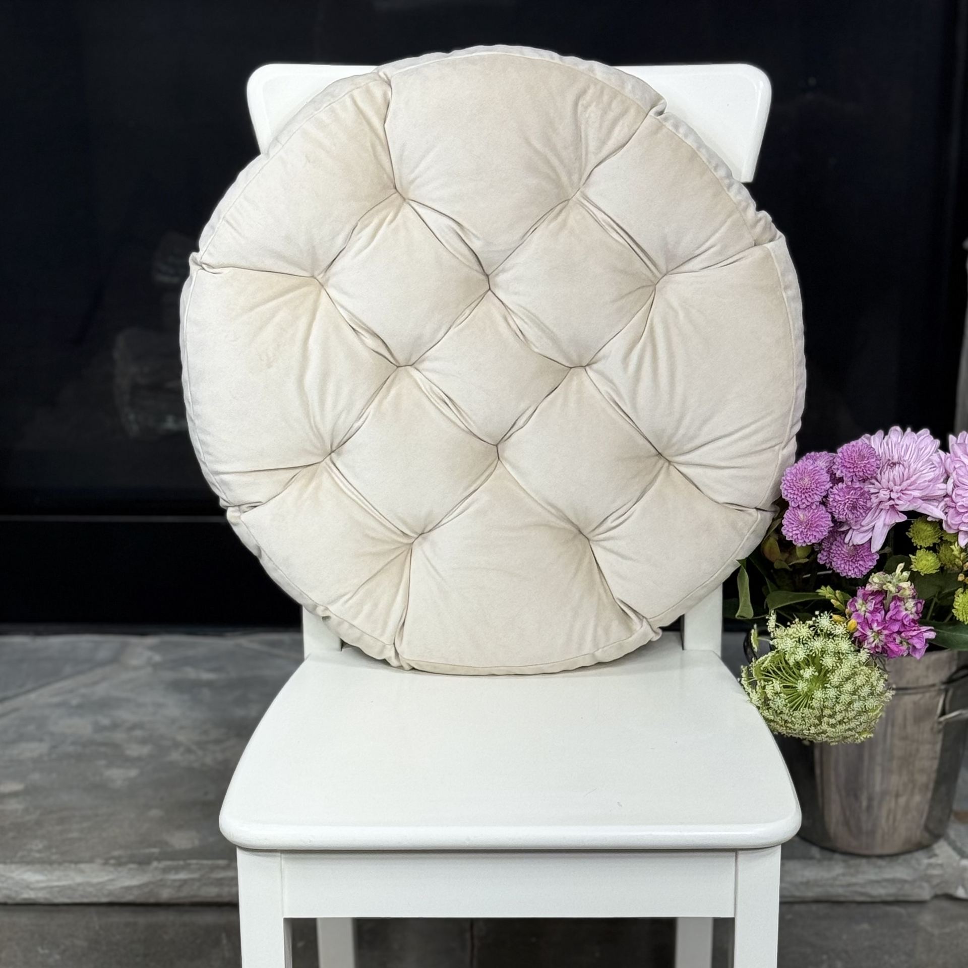 A white wooden dining chair with a large, round, tufted cream cushion on the back, styled in an indoor setting with a patterned rug and a vase of purple flowers.