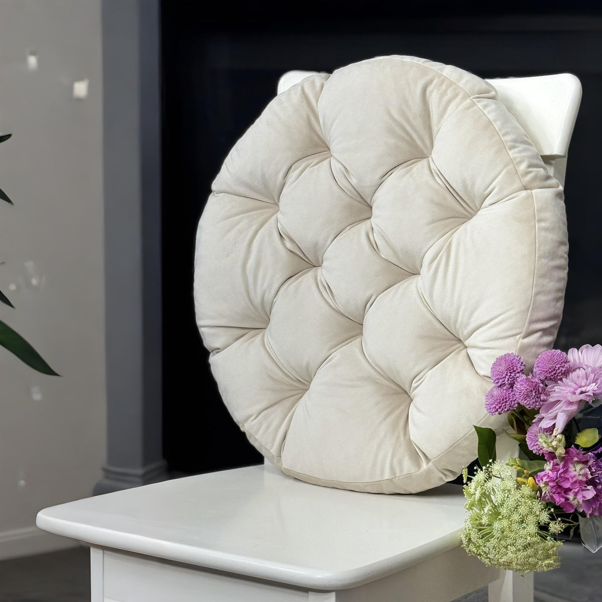 A white wooden dining chair with a large, round, tufted cream cushion on the back, styled in an indoor setting with a patterned rug and a vase of purple flowers.