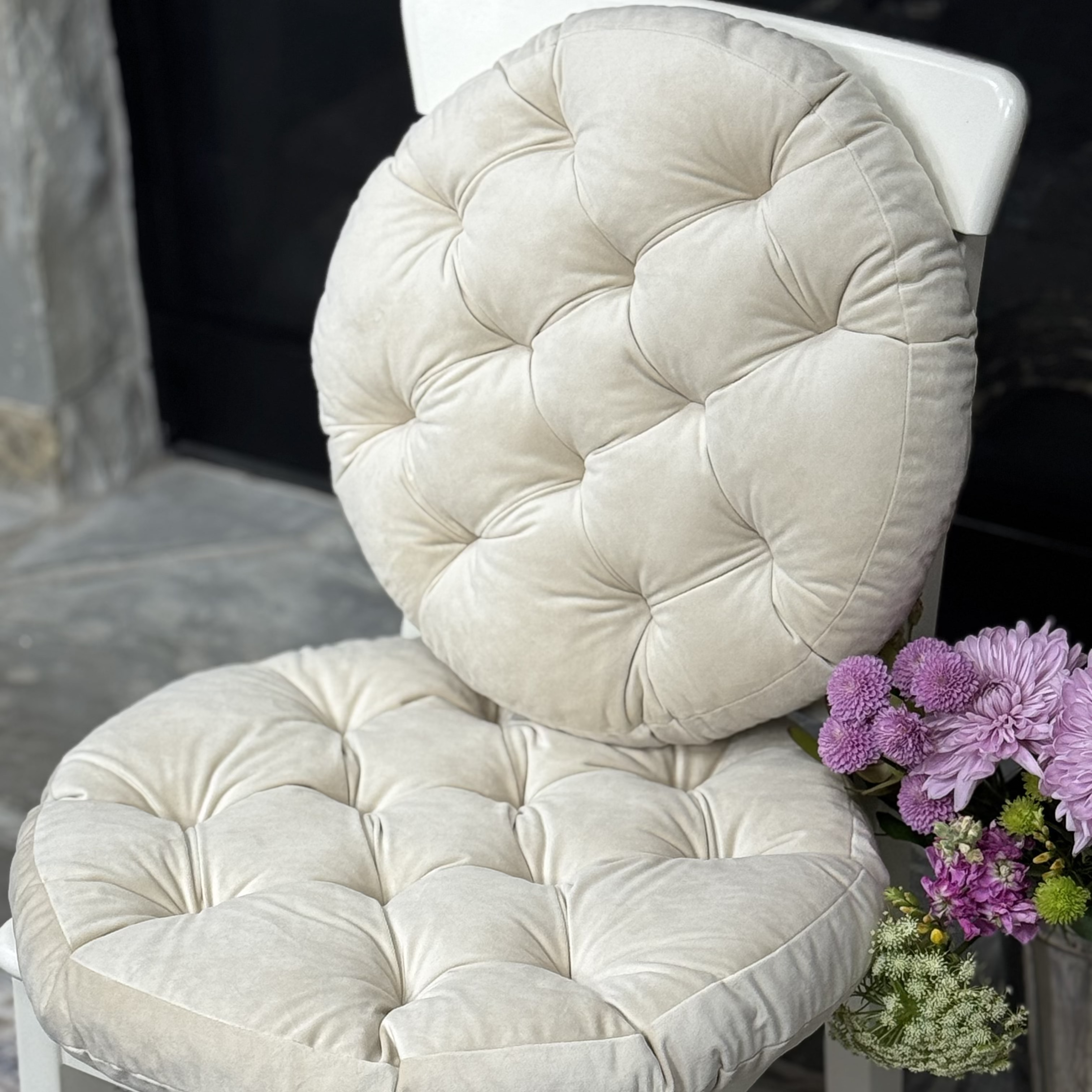 A white wooden dining chair with a large, round, tufted cream cushion on the back, styled in an indoor setting with a patterned rug and a vase of purple flowers.