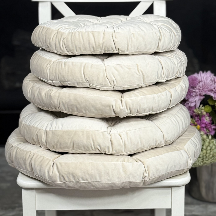 Four round tufted floor cushions stacked vertically on a white wooden chair, with a dark background and a vase of purple flowers on the right.